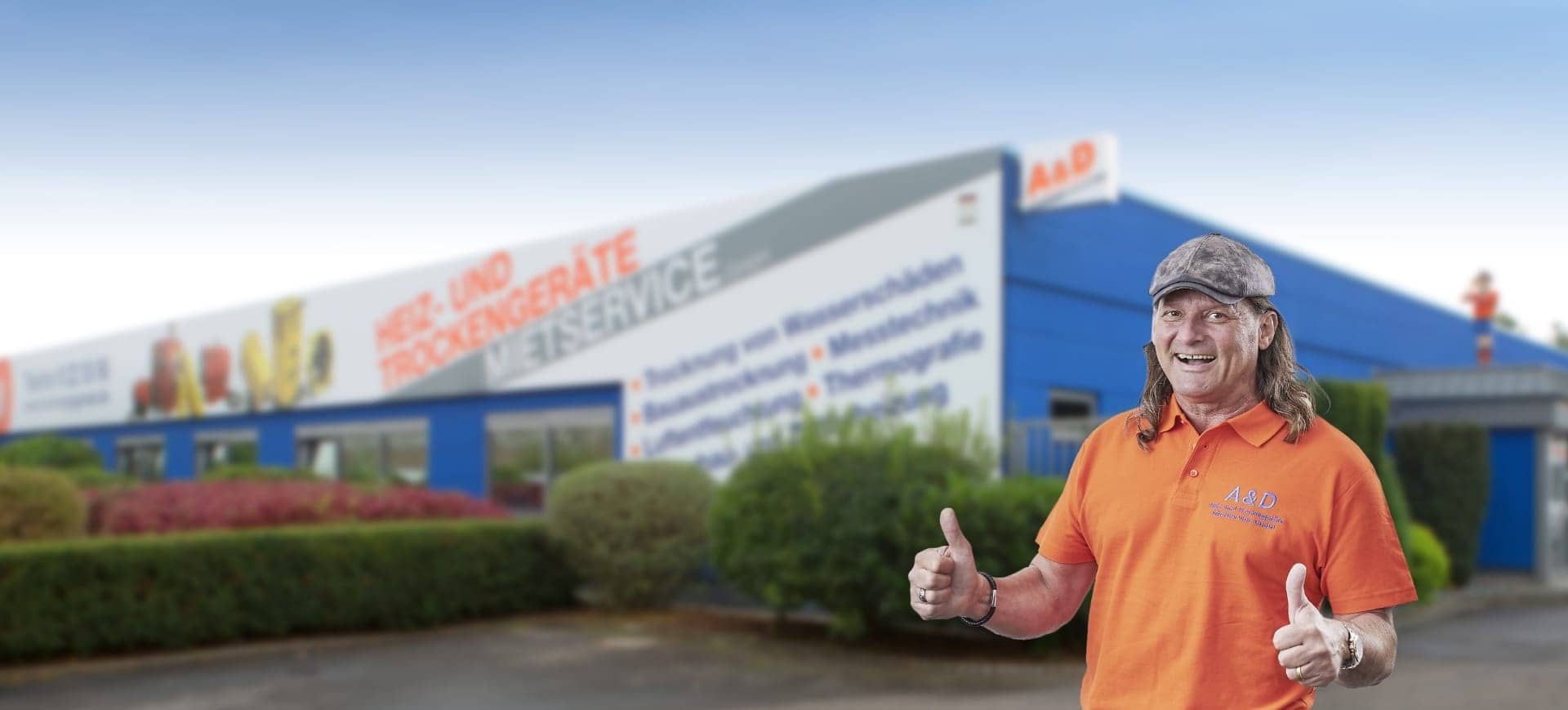 Albrecht Fischer, Geschäftsführer der A&D Heiz- und Trockengeräte Mietservice GmbH, steht vor dem Firmengebäude. Er trägt ein orangefarbenes Polohemd mit Logo und zeigt lächelnd beide Daumen nach oben.